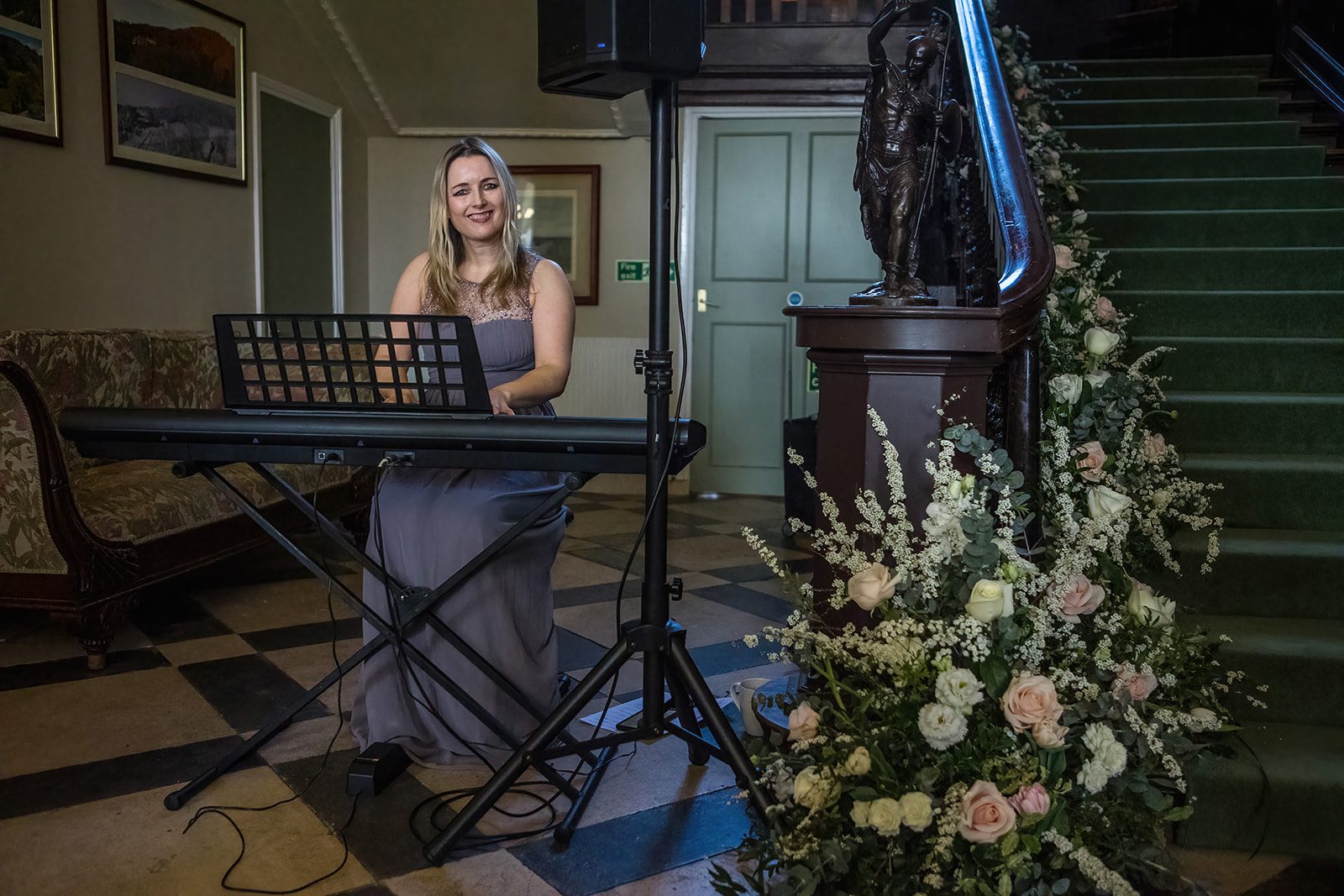 A woman playing piano next to the staircase at Crowcombe Court next to a floral decoration.